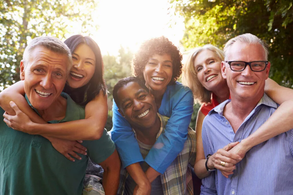 Group of six adults smiling together outdoors in sunlight with trees in background