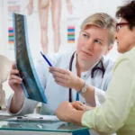 Female doctor in white coat showing spinal X-ray to patient while pointing with pen, with anatomical spine model on desk